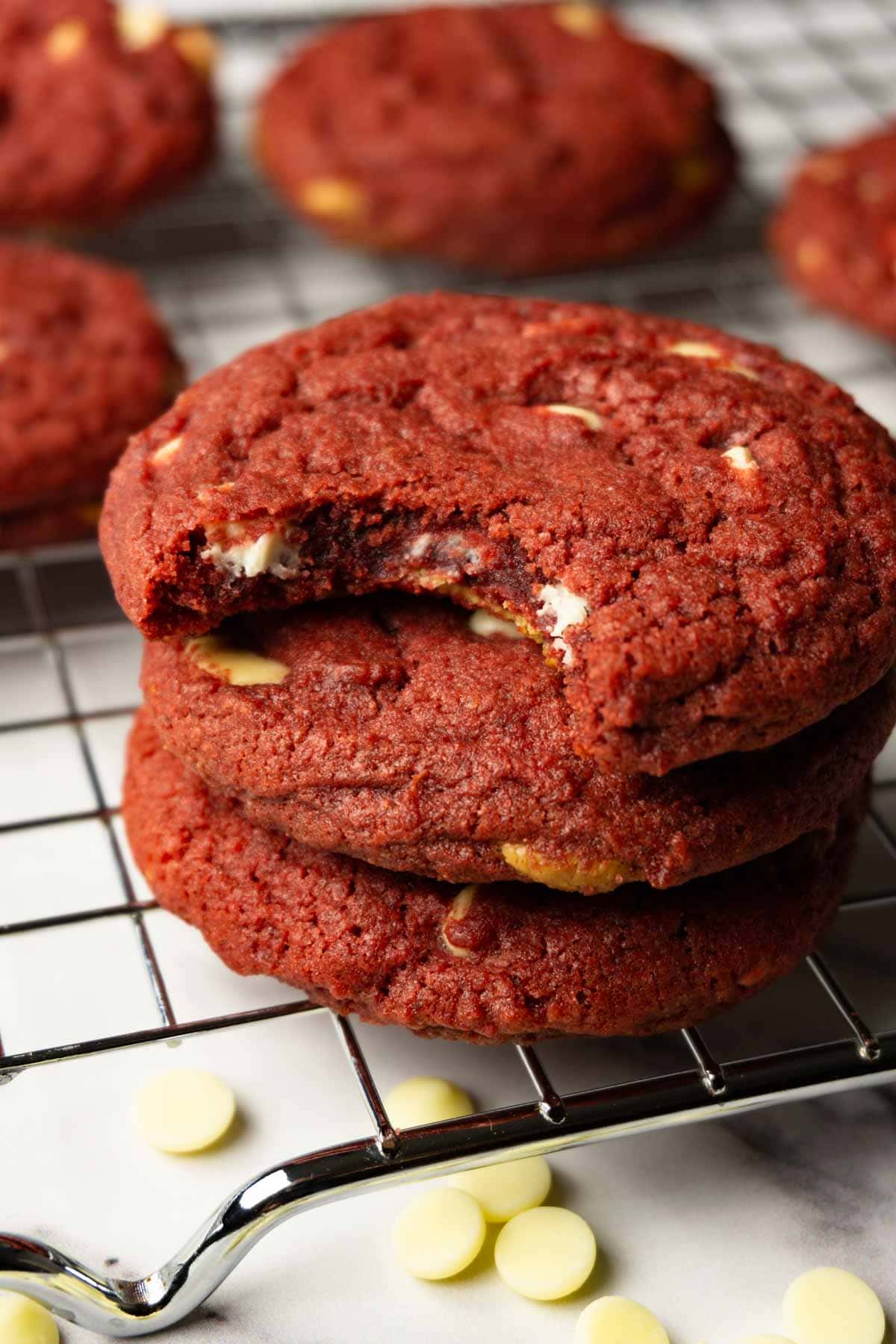 Three red velvet cookies with white chocolate chips stuck on top of each other, one bite taken from the top cookie.
