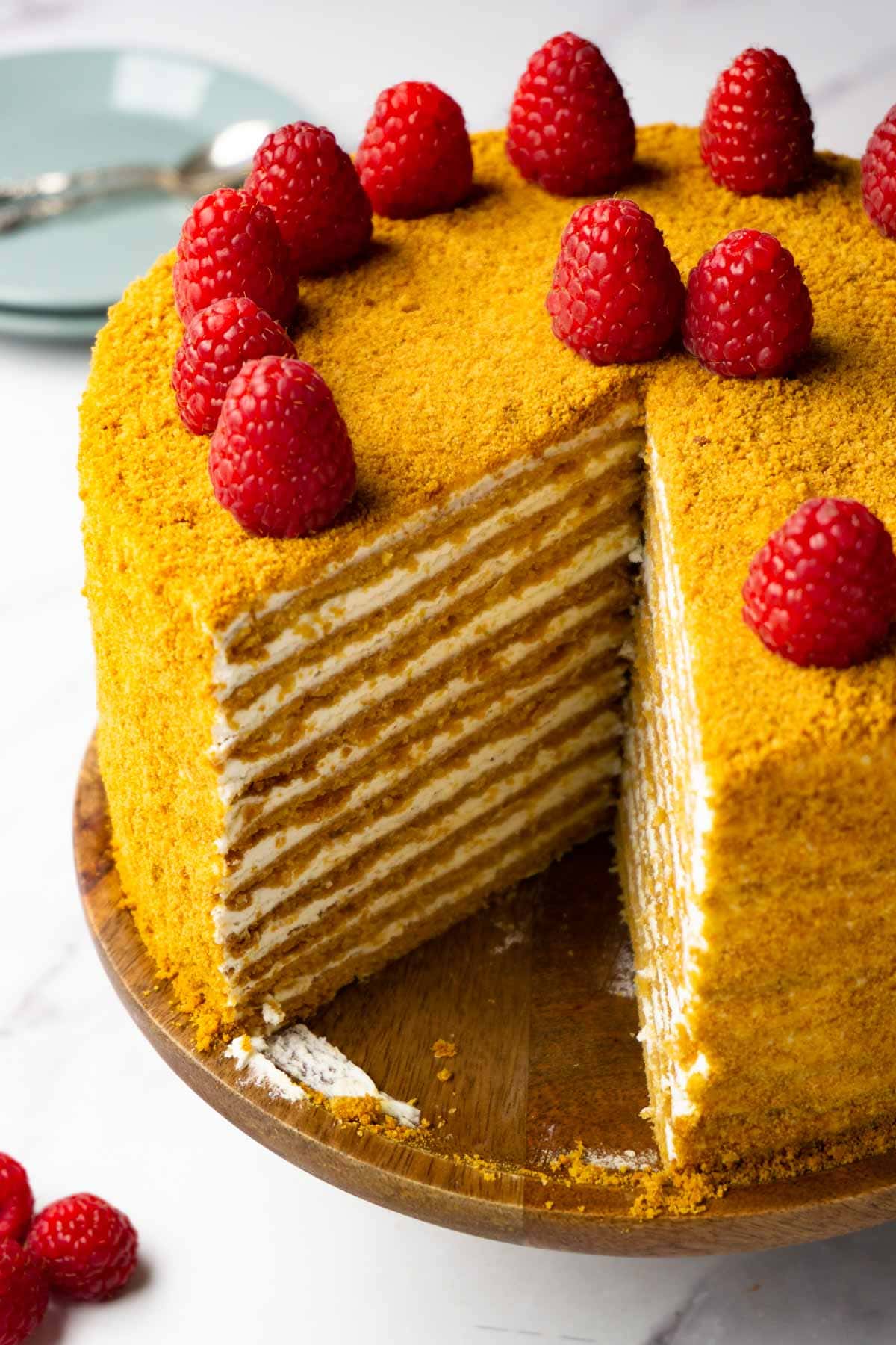 Honey cake decorated with fresh raspberries on a wooden cake stand, one piece taken.