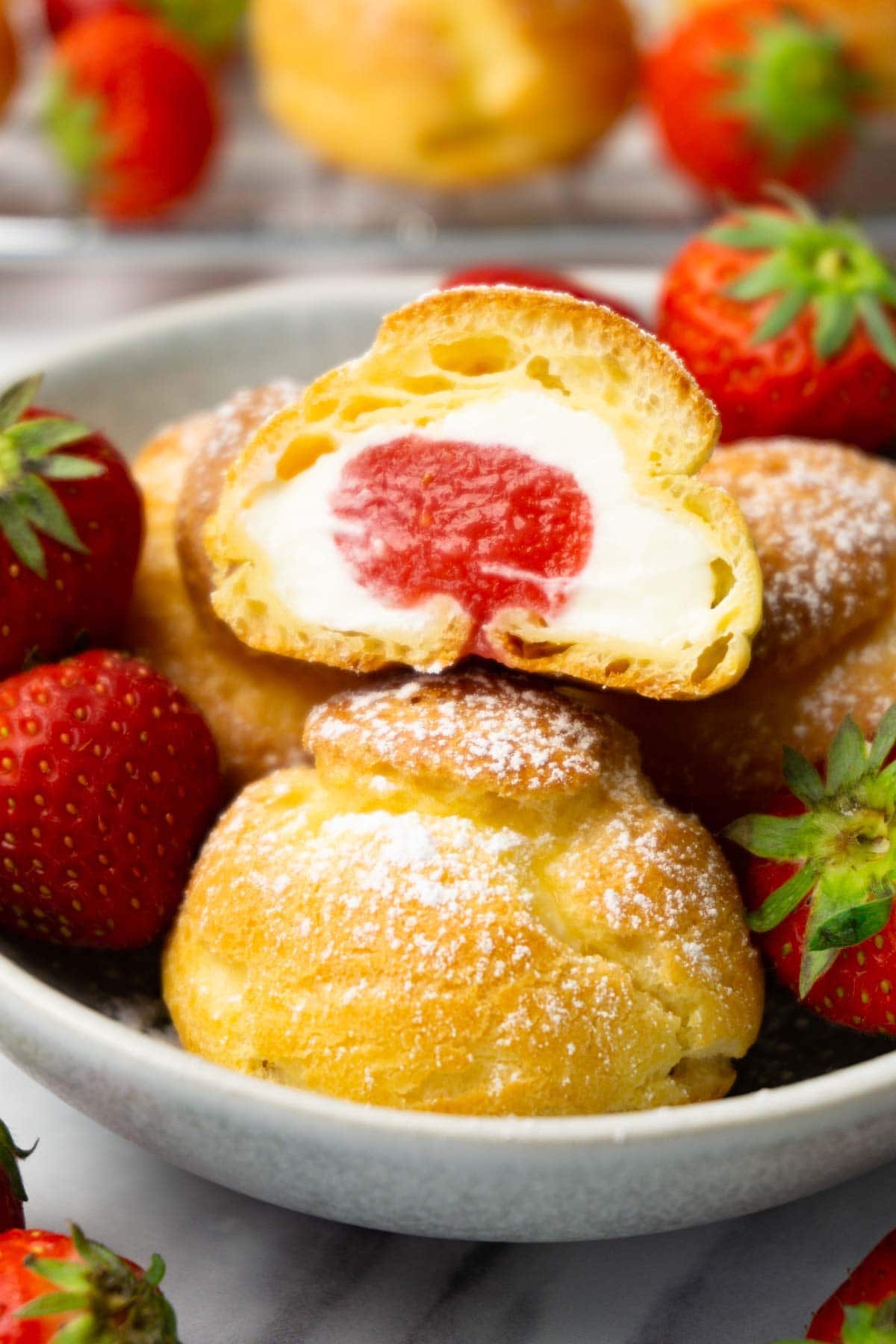 Cut in half cream puff filled with cream cheese filling and strawberry sauce in a bowl with more cream puffs and fresh strawberries.