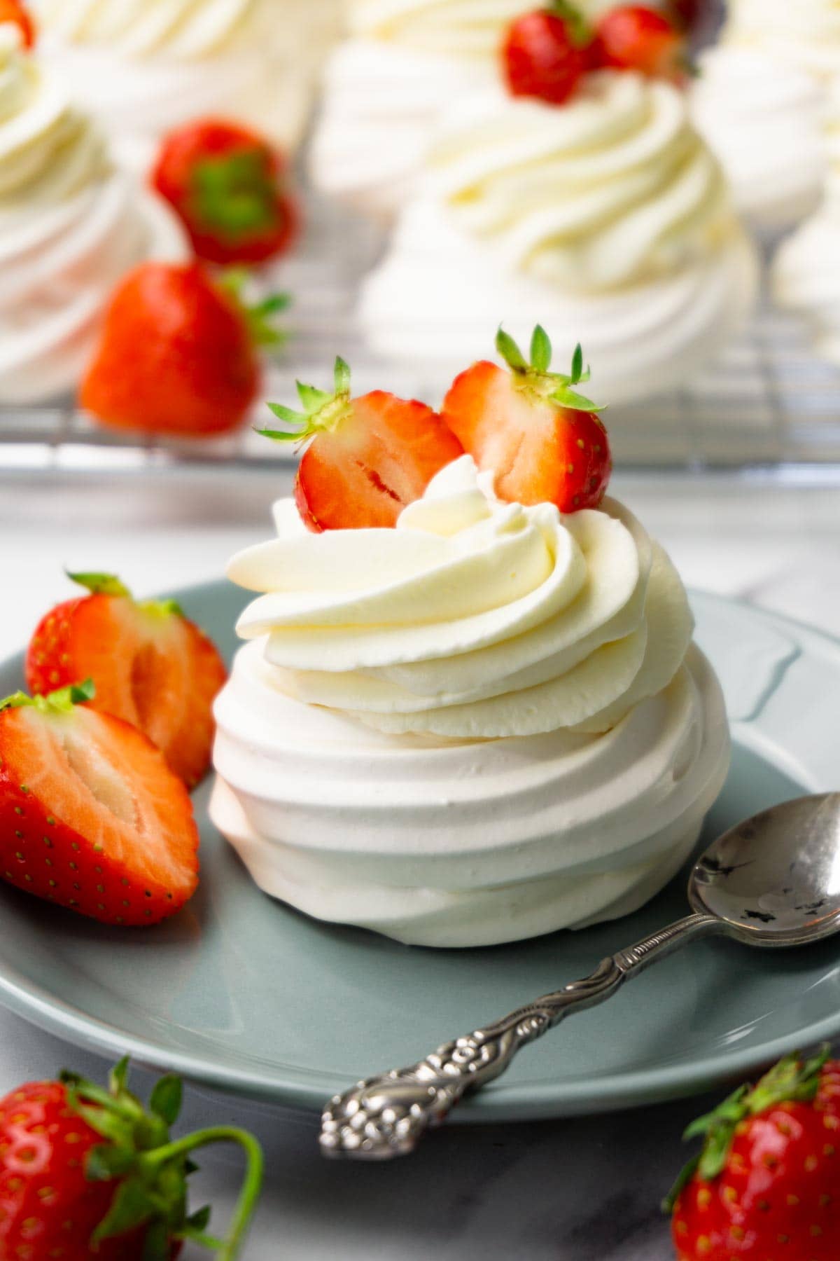Mini pavlova nest topped with cream cheese frosting and strawberries on a small round plate, more pavlovas on a cooling rack on the background.