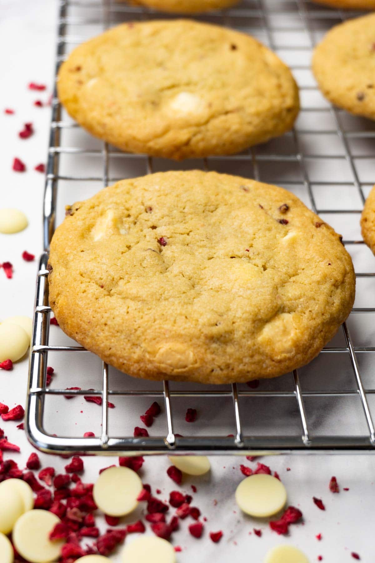 A white chocolate cookies on a cooling rack, white chocolate chips and freeze dried raspberries are lying next to it.