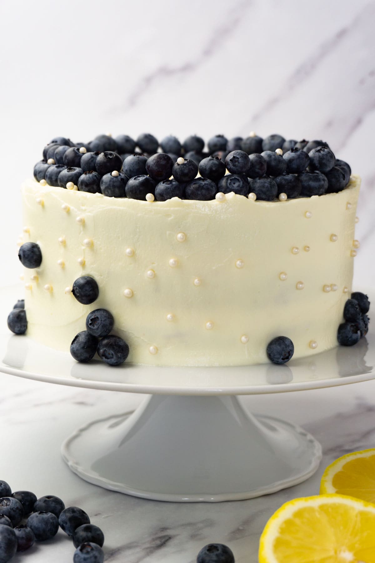 Lemon curd cake on a white cake server frosted with cream cheese frosting and decorated with sugar purls and fresh blueberries.