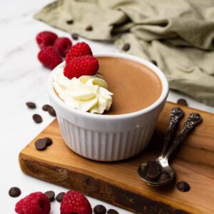 Dark chocolate mousse topped whipped cream and fresh raspberries in a white ramekin.