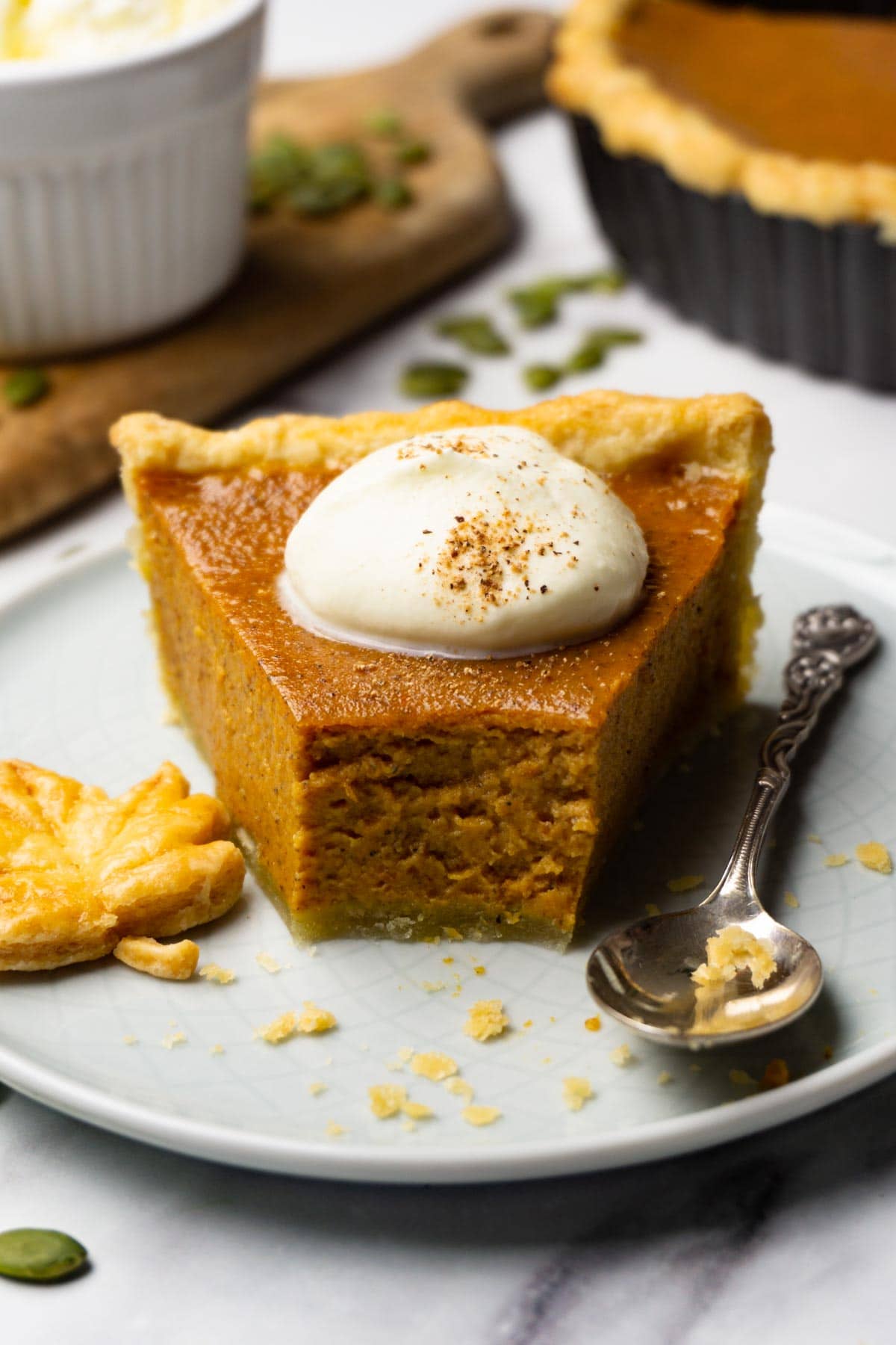 A slice of pumpkin pie topped with whipped heavy cream and ground nutmeg on a small round plate with silver spoon and a maple leave made of puff pastry, one bite taken.