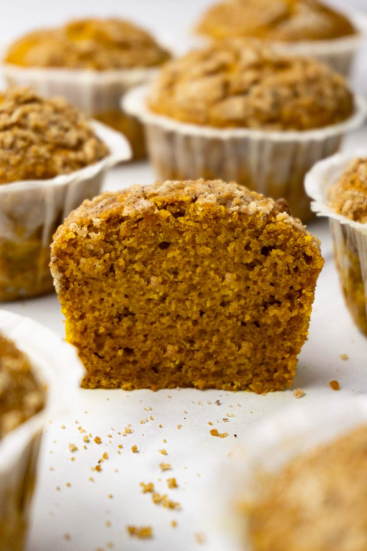 Half of pumpkin muffin with streusel topping on a white surface, more muffins are lying around.