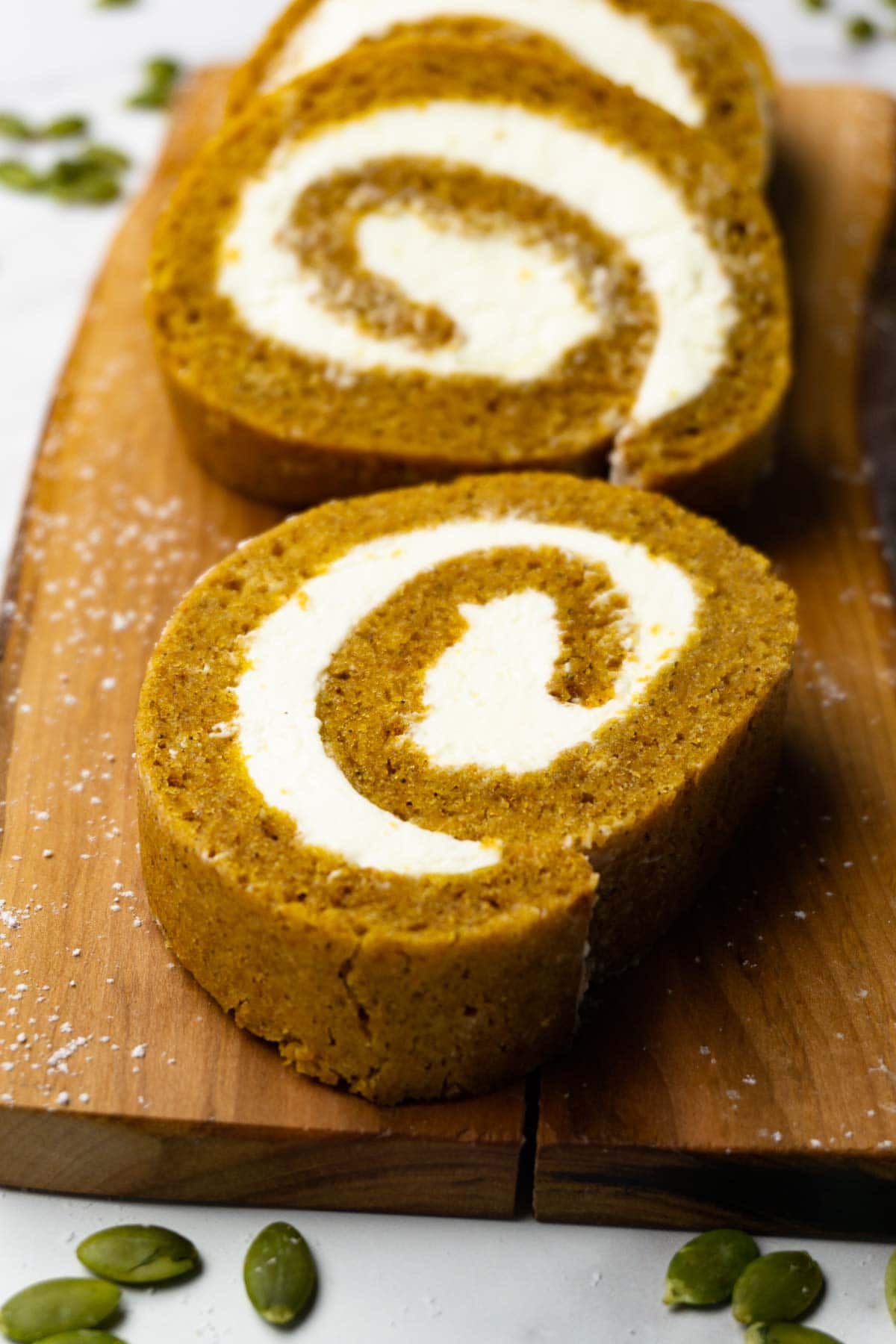 Slices of pumpkin roll with cream cheese frosting on a wooden board, pumpkin seeds are lying around.