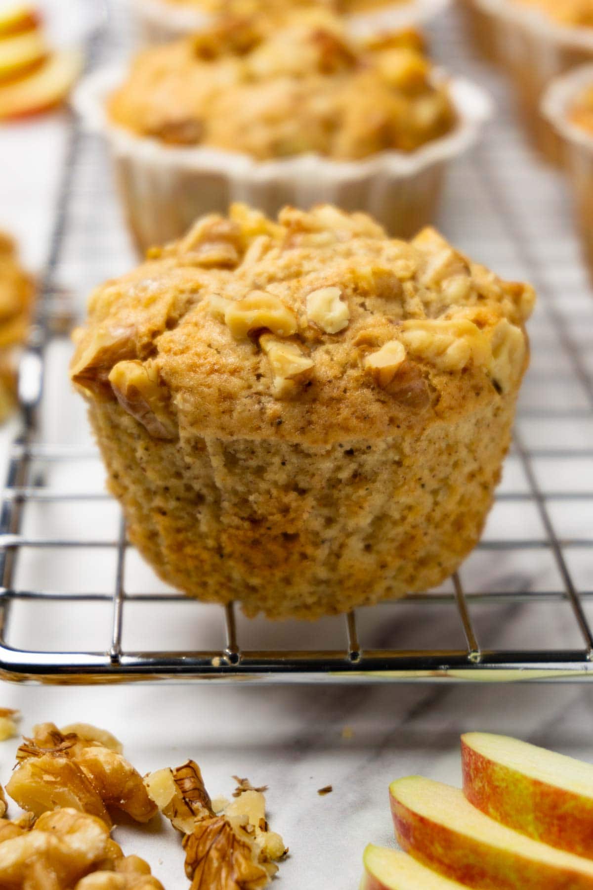 Apple muffins topped with crushed walnuts on a cooling rack, muffin line removed from one muffin.