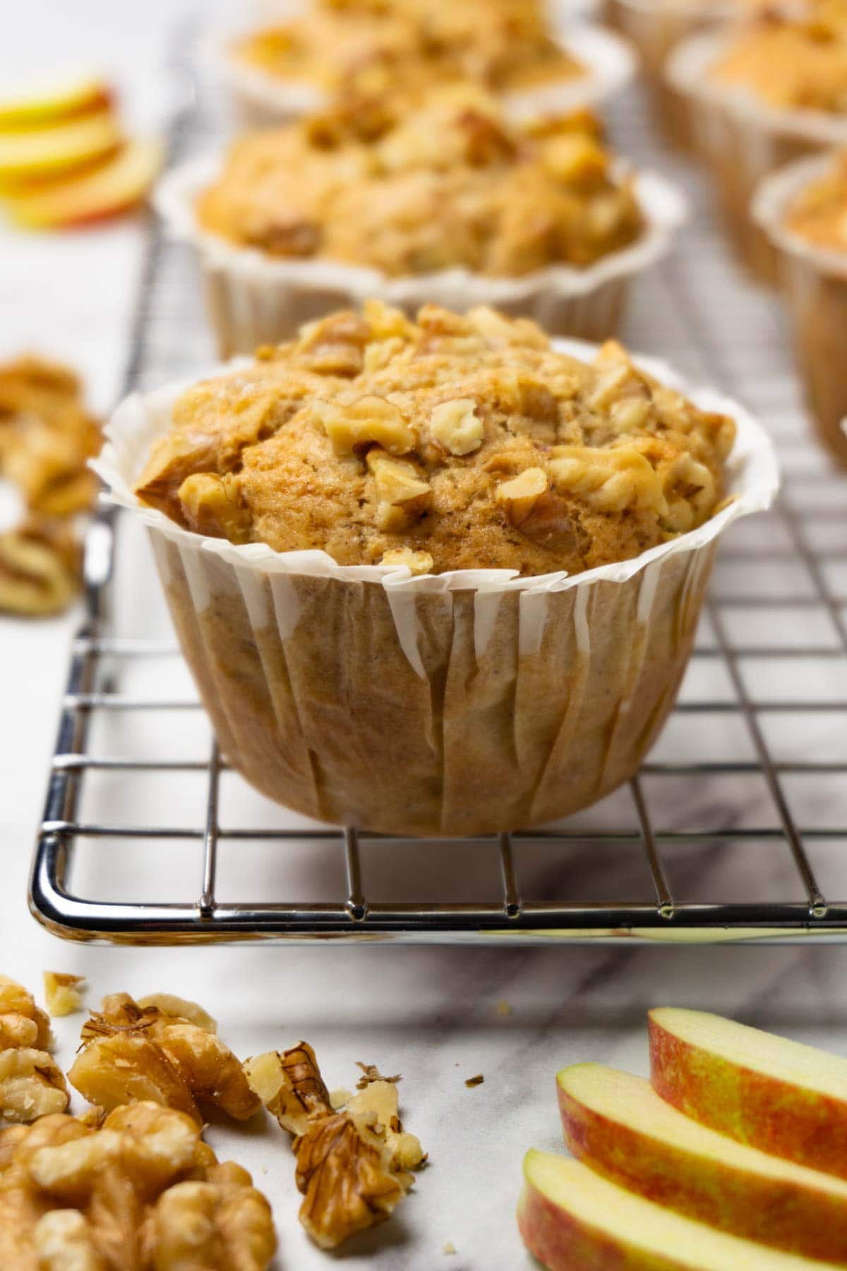 Apple muffins topped with crushed walnuts on a cooling rack, fresh apples and more walnuts are lying around.