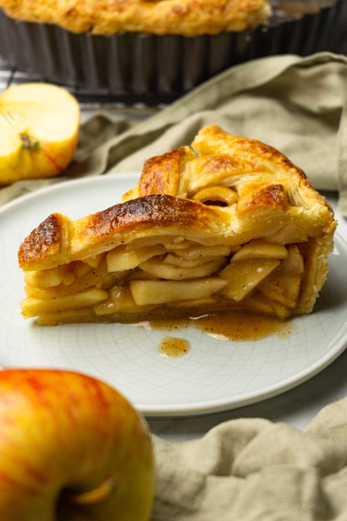 A piece of apple pie served on a small round plate, fresh apples are lying around.