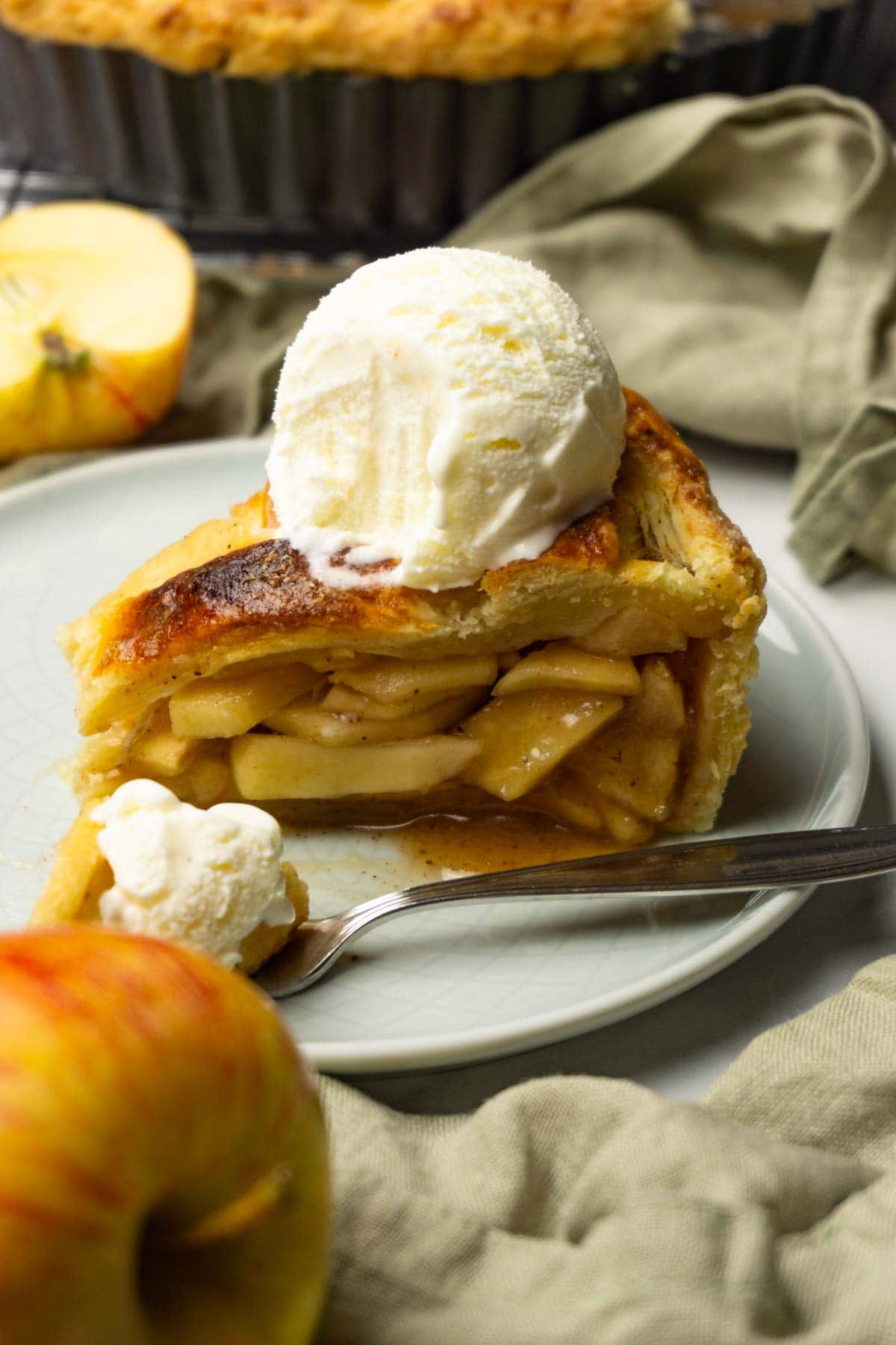 A piece of apple pie with a scoop of vanilla ice cream on top served on a small round plate, one bite taken from it, fresh apples are lying around.