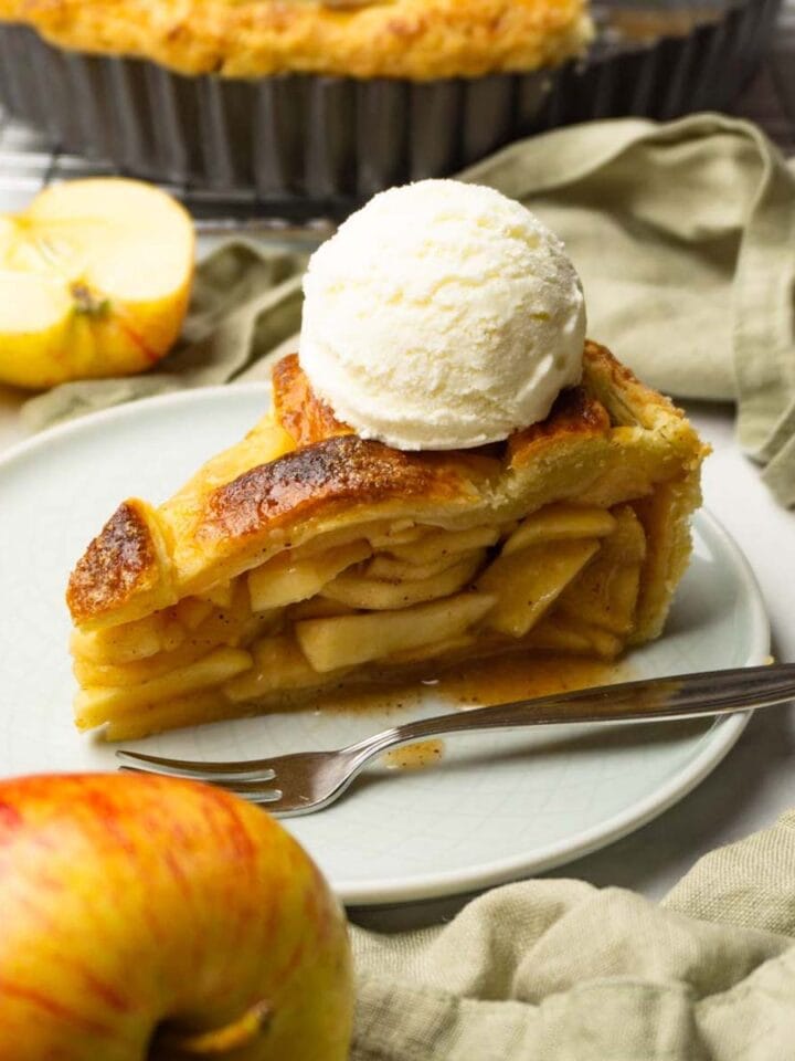 A piece of apple pie with a scoop of vanilla ice cream on top served on a small round plate, fresh apples are lying around.