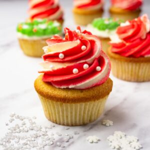 A cupcake decorated with two-tone (red and white) cream cheese frosting and pearl-like sprinkles, more cupcakes on the background.
