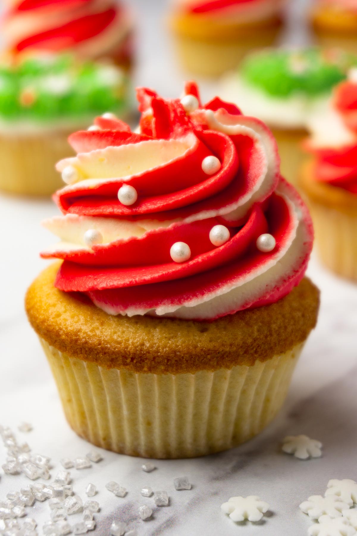 A cupcake decorated with two-tone (red and white) cream cheese frosting and pearl-like sprinkles, more cupcakes on the background.