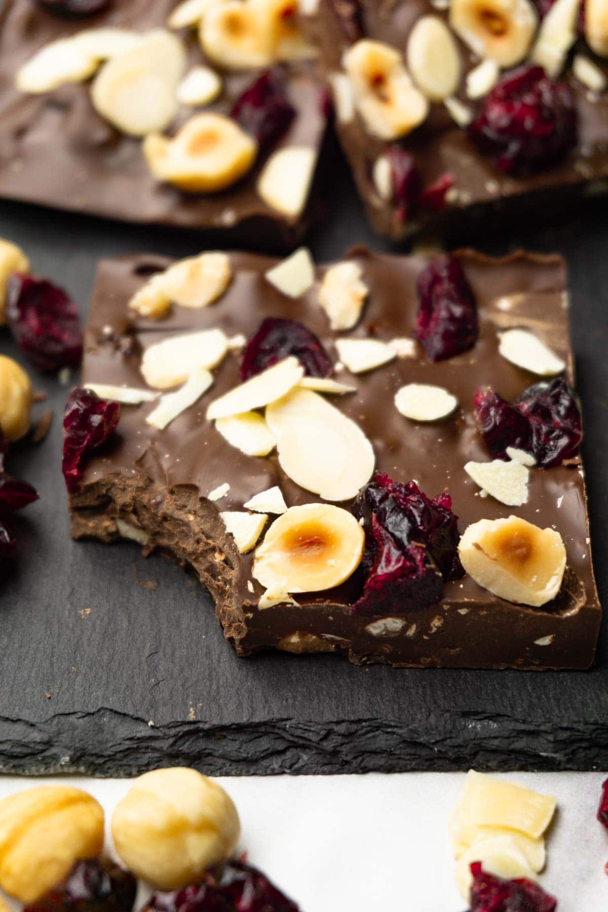 Pieces of dark chocolate bark with dried cranberries and roasted hazelnuts served on a dark serving stone board, one bite taken from one of the pieces.