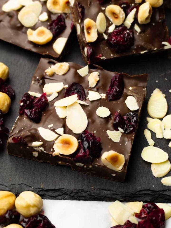 Pieces of dark chocolate bark with dried cranberries and roasted hazelnuts served on a dark serving stone board.