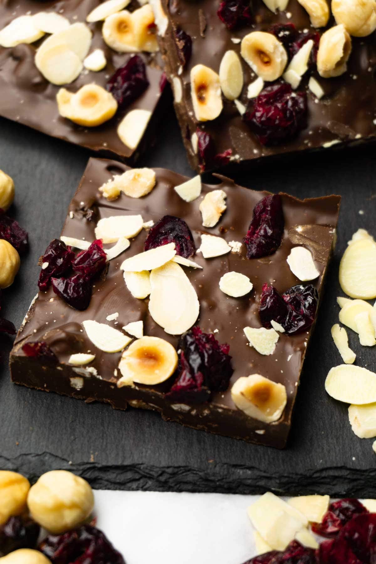 Pieces of dark chocolate bark with dried cranberries and roasted hazelnuts served on a dark serving stone board.