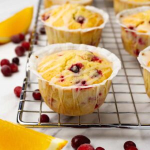 Cranberry orange muffins with sugar glaze are on a metal cooling rack. Fresh orange slices and cranberries are lying around.
