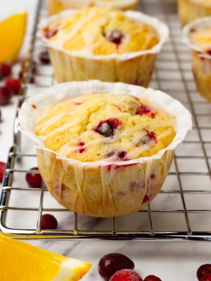 Cranberry orange muffins with sugar glaze are on a metal cooling rack. Fresh orange slices and cranberries are lying around.