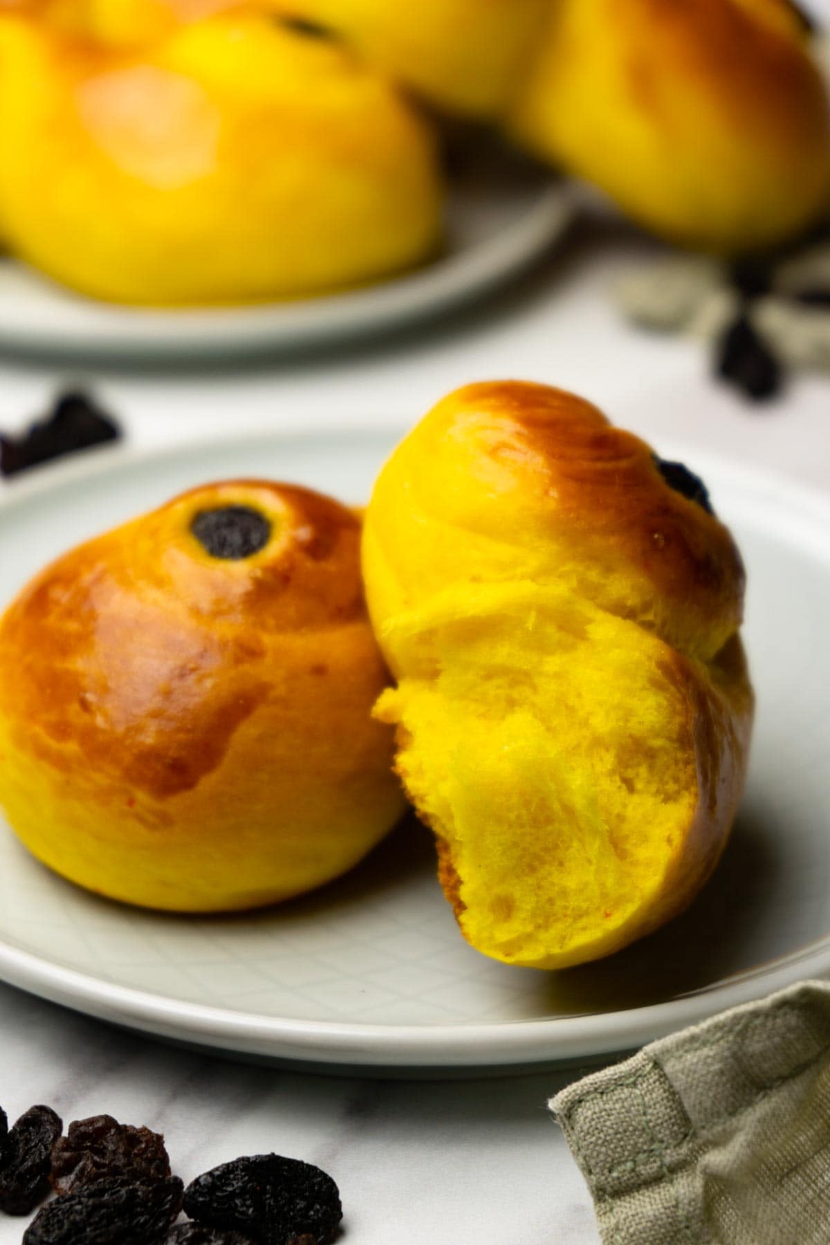 Broken in half saffron bun with raisins on a small round plate, more buns on the background.