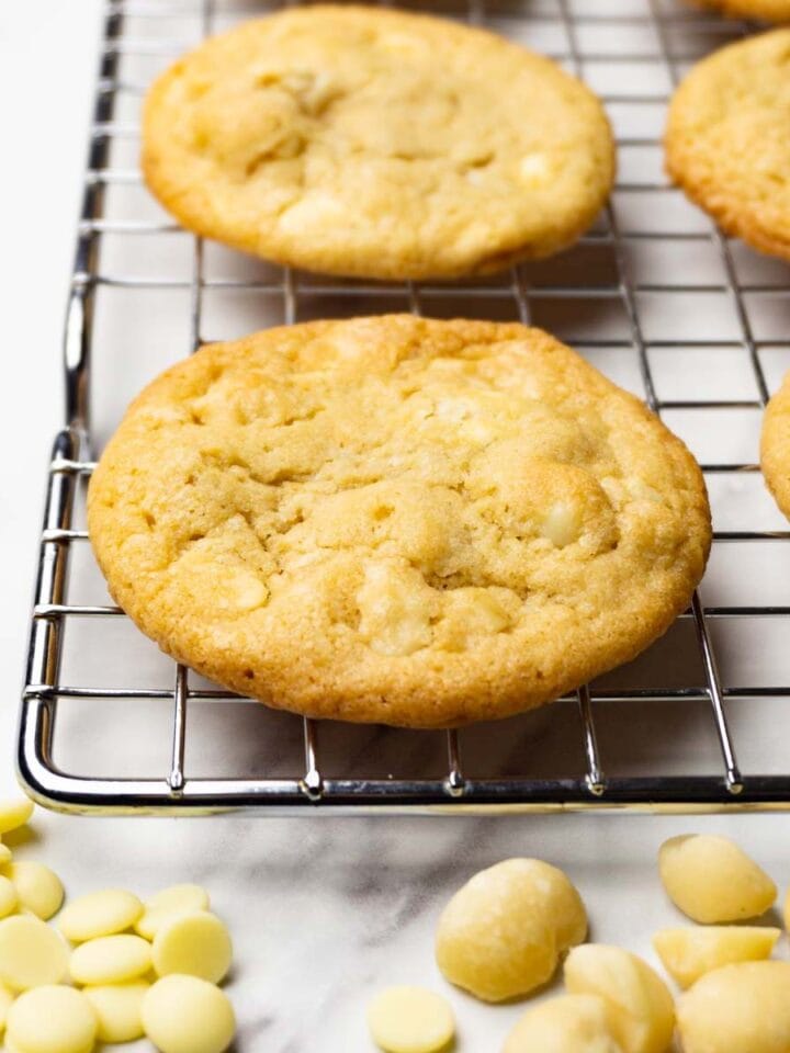 Cookies with white chocolate and macadamia nuts on a metal rack, white chocolate chips and macadamia nuts are lying around.