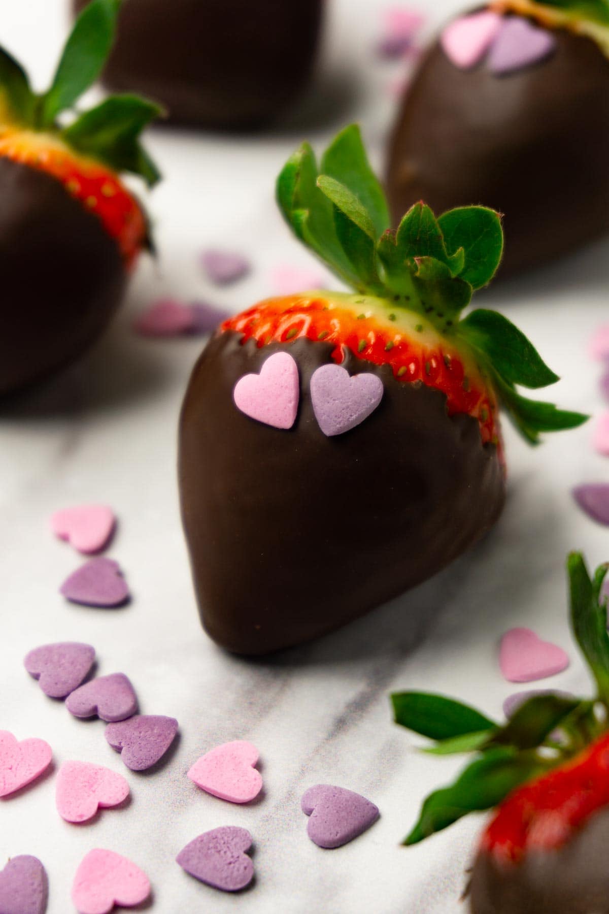 Dark chocolate-covered strawberries with heart-shaped sprinkles on a marble working surface.