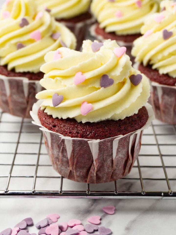 Red velvet cupcakes frosted with cream cheese frosting and decorated with heart-shaped sprinkles.