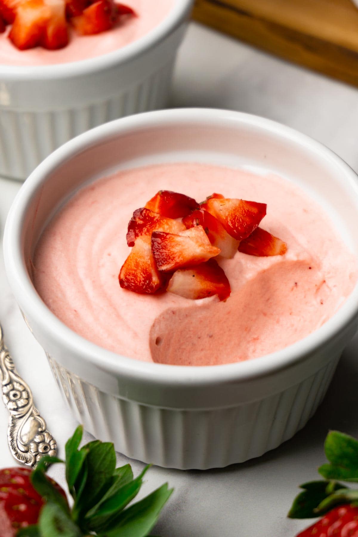 Strawberry mousse decorated with cubed fresh strawberries served in a white ramekin, one spoon is taken.