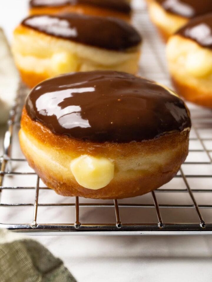 Donuts filled with pastry cream and glazed with dark chocolate ganache on a metal cooling rack.
