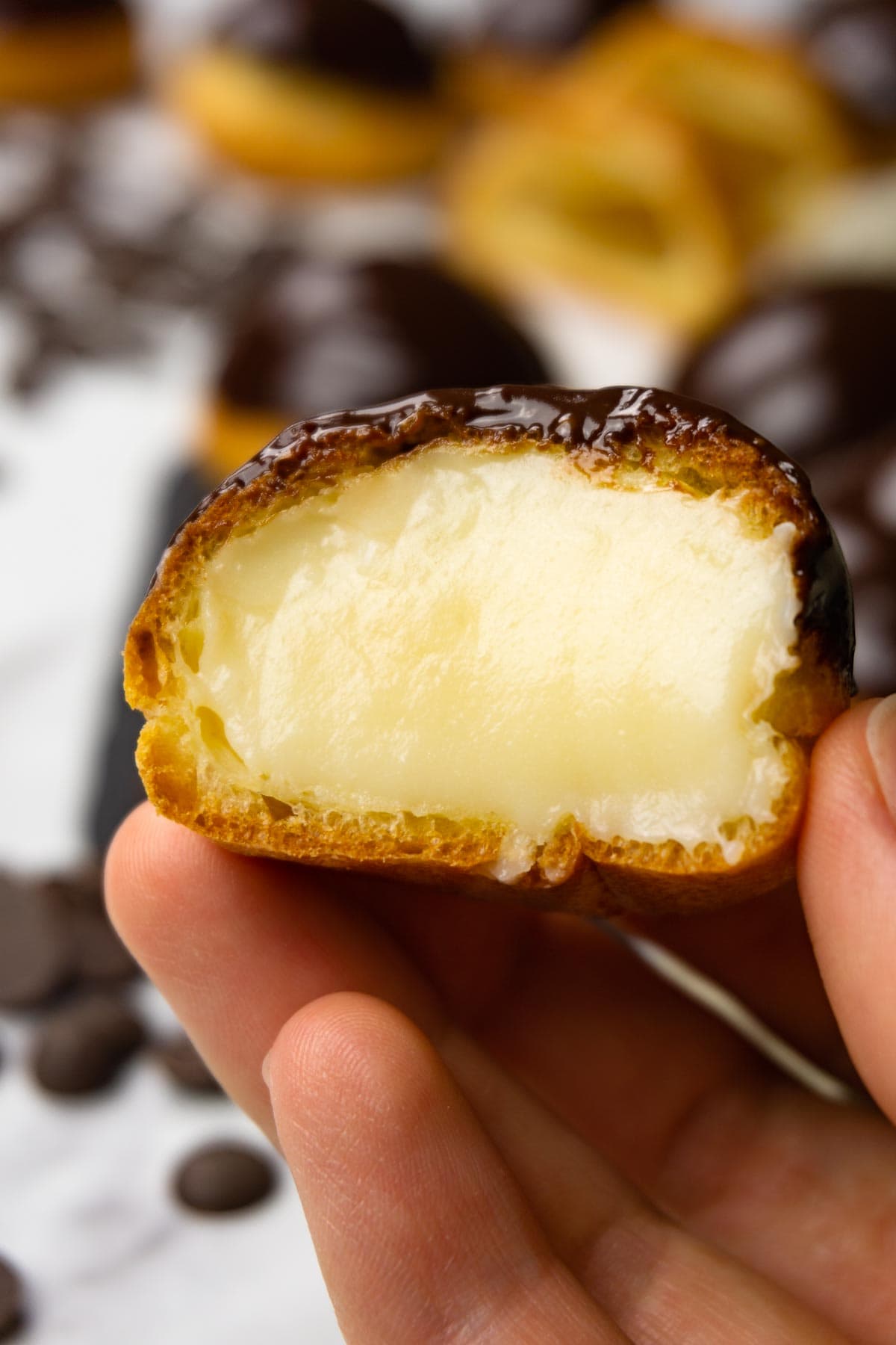 A hand is holding a profiterole filled with pastry cream and glazed with chocolate ganache, one bite is taken.