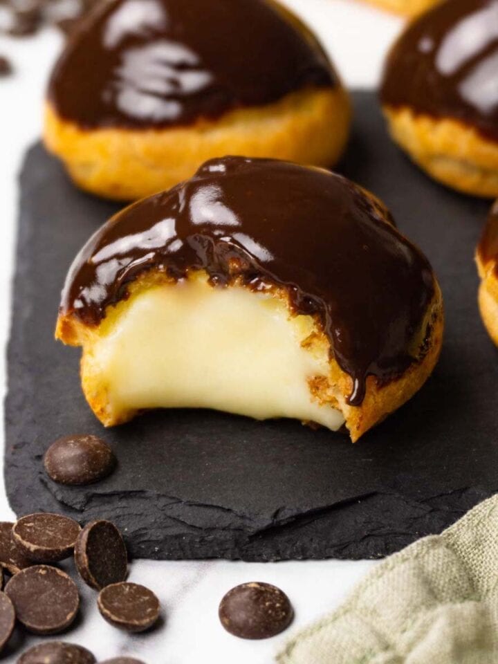 Profiteroles filled with pastry cream and glazed with chocolate ganache on a dark stone serving board, one bite is taken from one of the profiteroles.