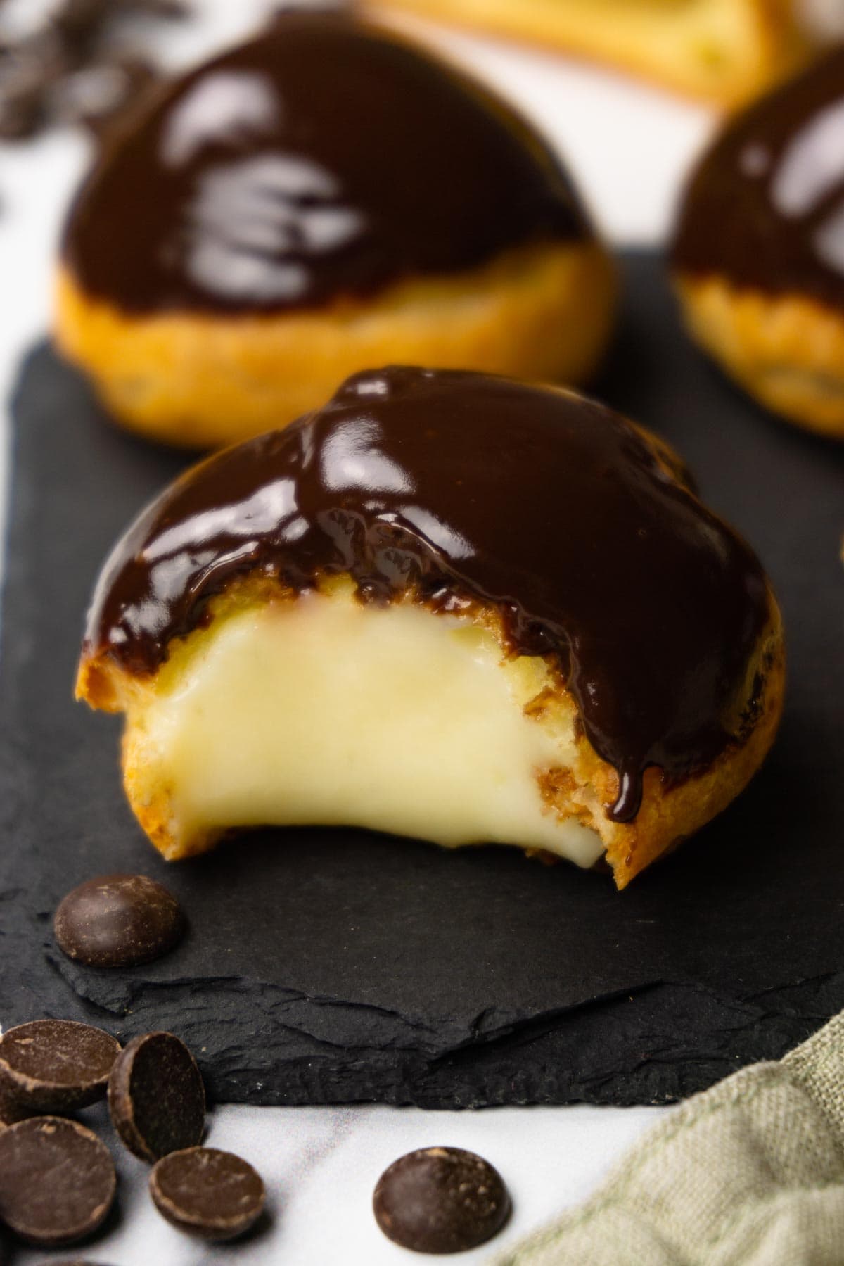 Profiteroles filled with pastry cream and glazed with chocolate ganache on a dark stone serving board, one bite is taken from one of the profiteroles.