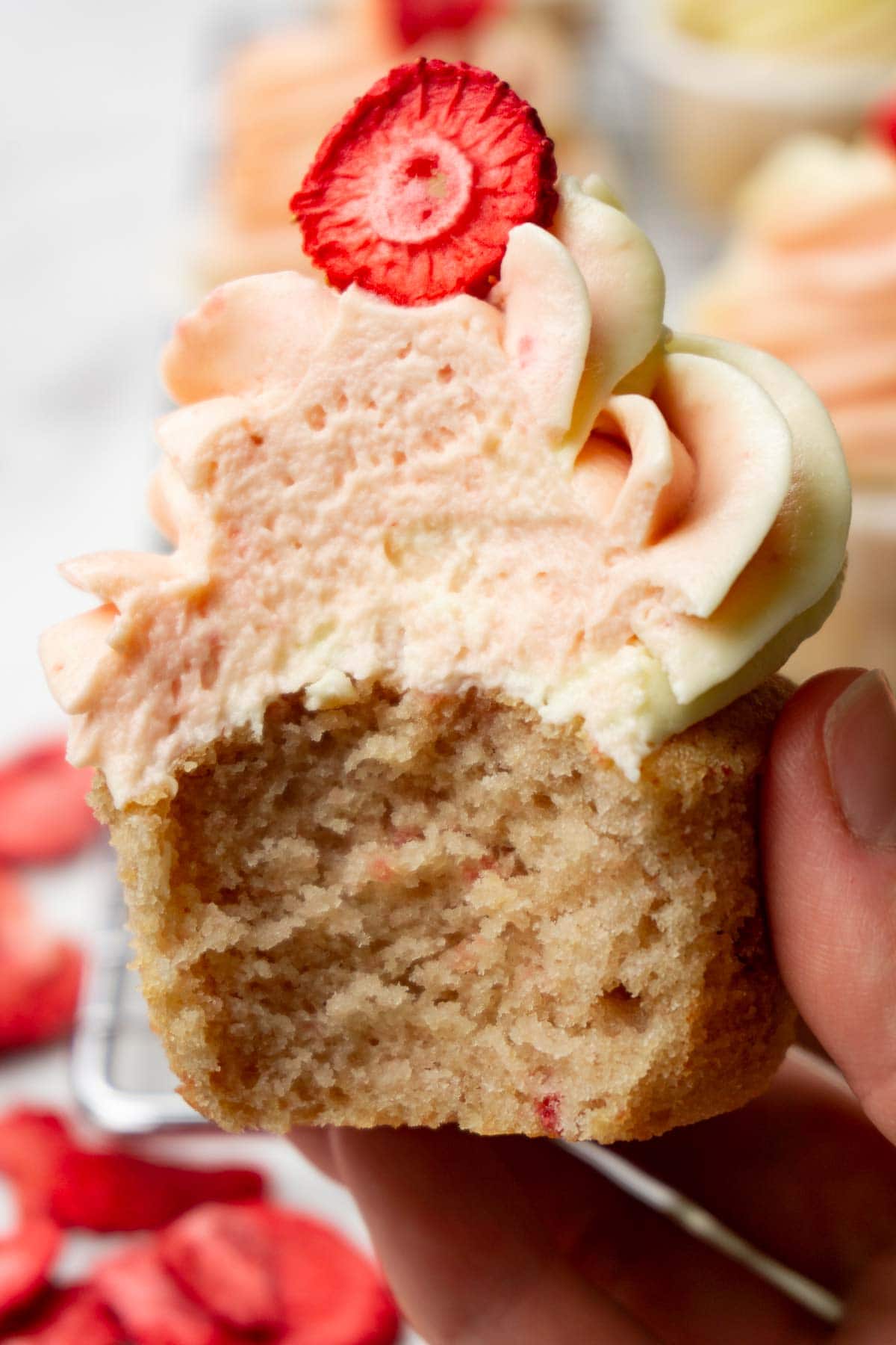 A hand is holding a strawberry cupcake with cream cheese frosting, one bite is taken.