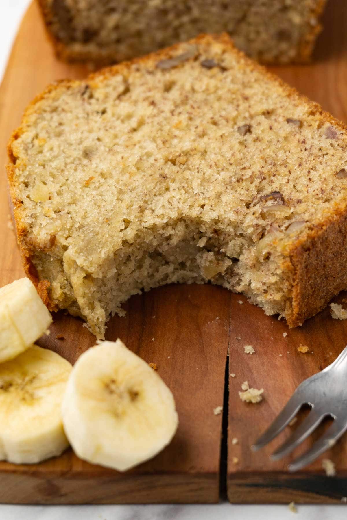 A slice of banana bread with walnuts on a wooden board; one bite was taken.