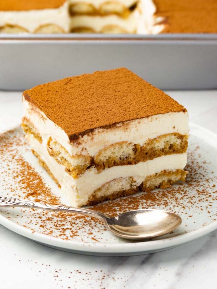 A piece of tiramisu dusted with cocoa powder on a round plate with a silver spoon.