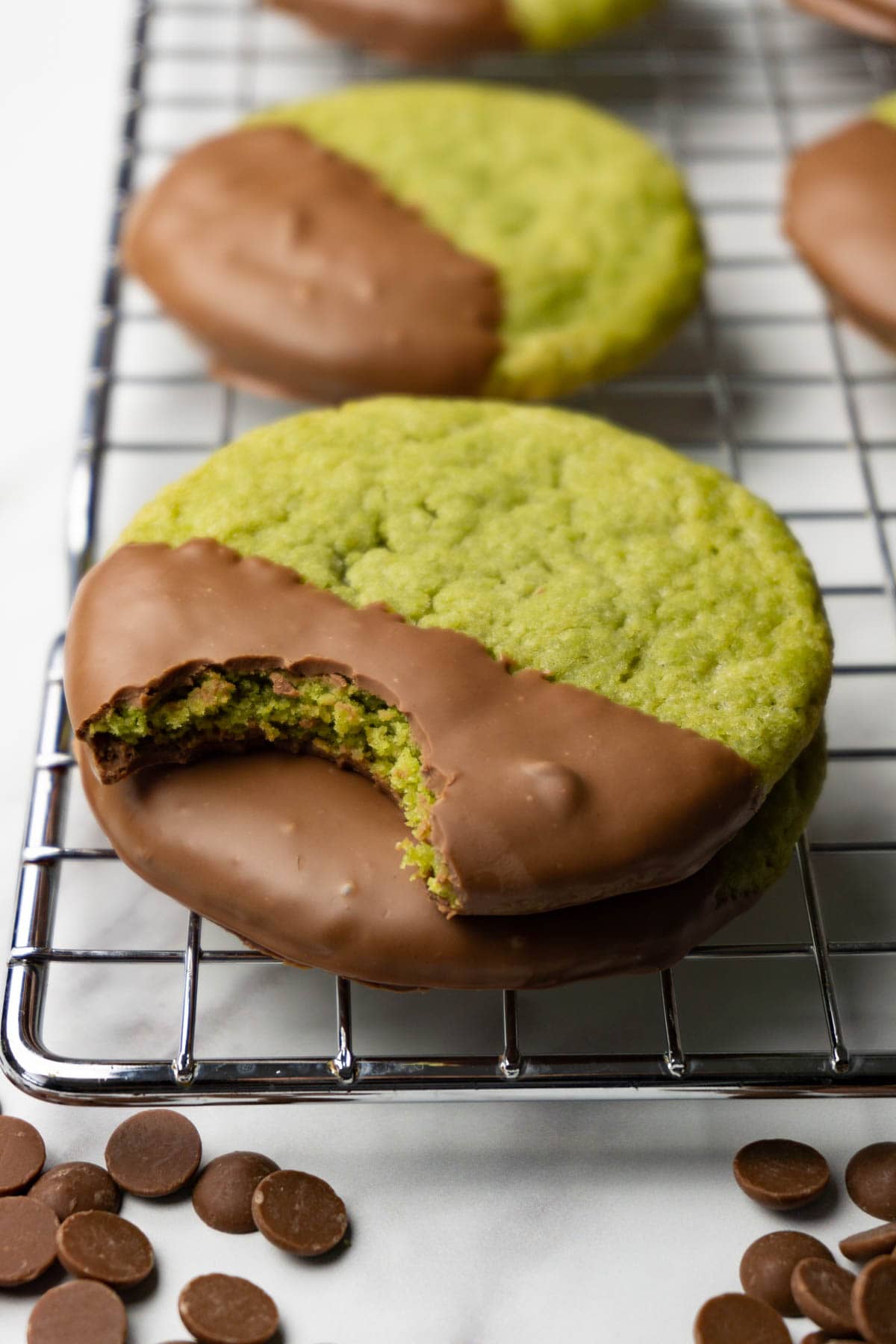 Matcha cookies with milk chocolate glaze stacked on top of each other on a cooling rack. One bite is taken from the top cookie.
