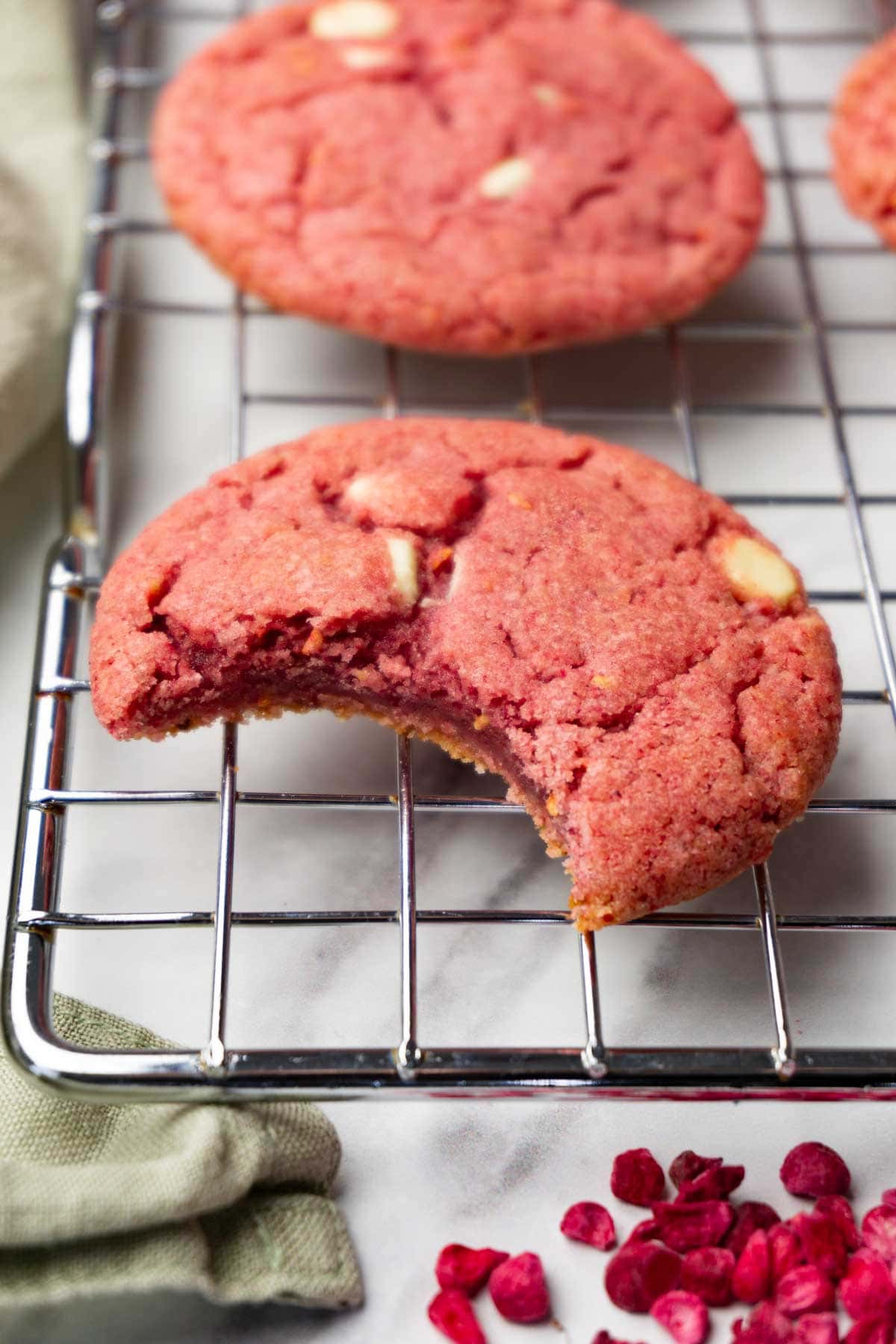 Raspberry cookies with white chocolate chips are lying on a wire rack; one bite was taken from one of the cookies.