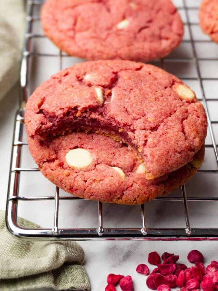Raspberry cookies with white chocolate chips are stacked on top of each other; one bite was taken from the top cookie.