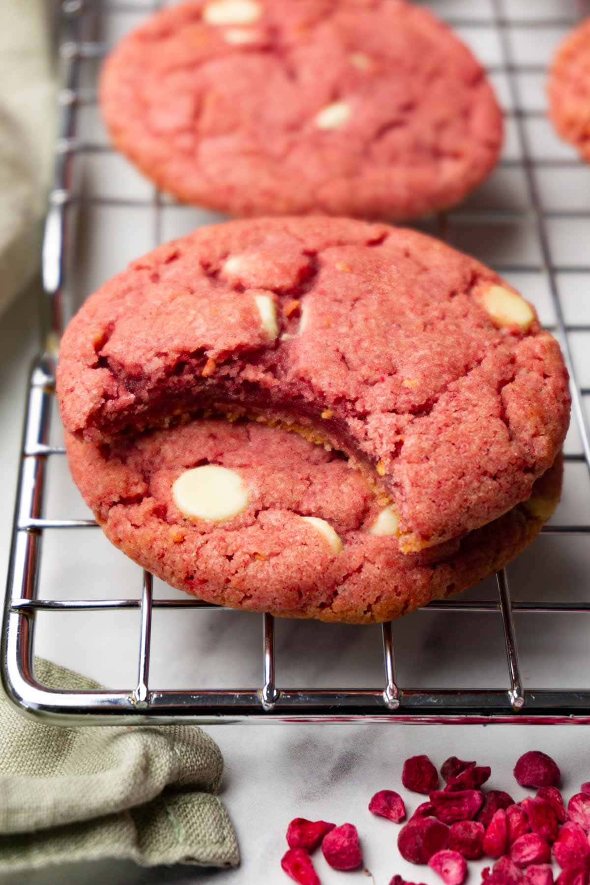 Raspberry cookies with white chocolate chips are stacked on top of each other; one bite was taken from the top cookie.