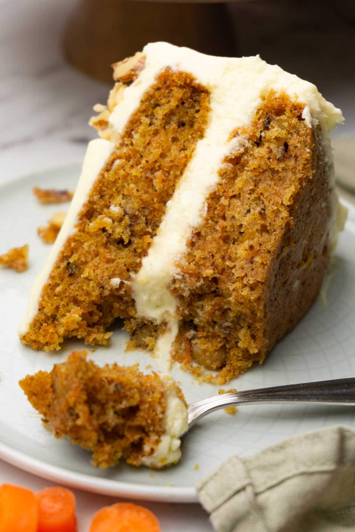 A piece of carrot cake with cream cheese frosting is decorated with chopped walnuts and served on a small round plate; one bite was taken.