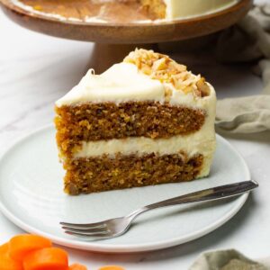 A piece of carrot cake with cream cheese frosting is decorated with chopped walnuts and served on a small round plate with a silver dessert fork.