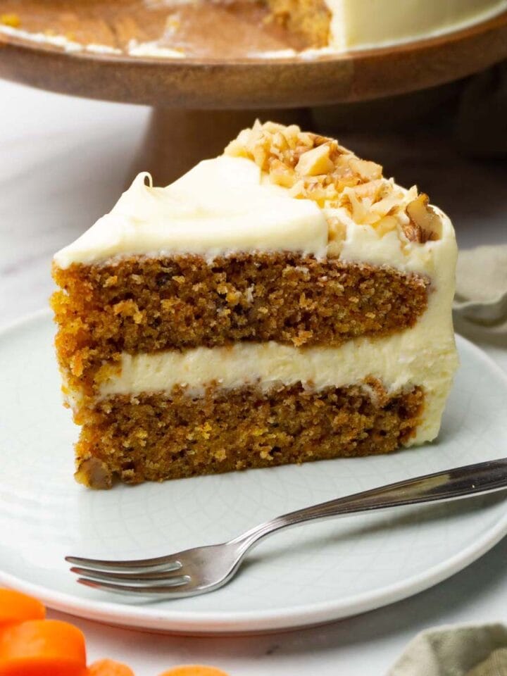 A piece of carrot cake with cream cheese frosting is decorated with chopped walnuts and served on a small round plate with a silver dessert fork.