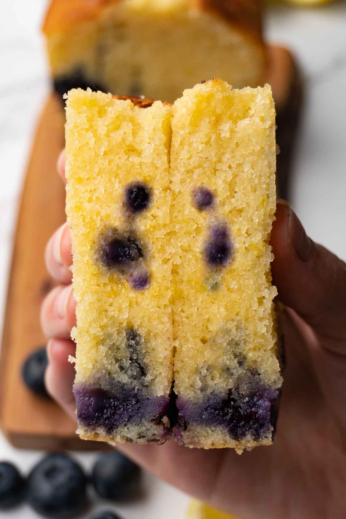 A hand is holding a cut in half slice of a pound cake with lemon zest and blueberries.