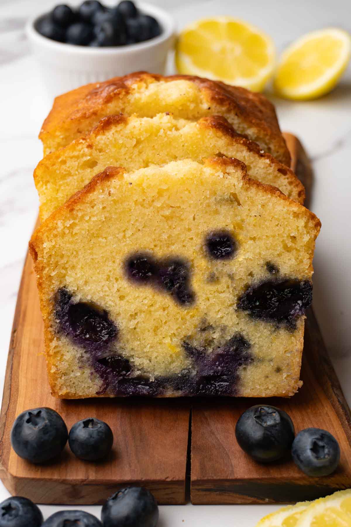 A sliced pound cake with lemon zest and blueberries on a wooden serving boars, fresh blueberries and lemon slices are lying around.