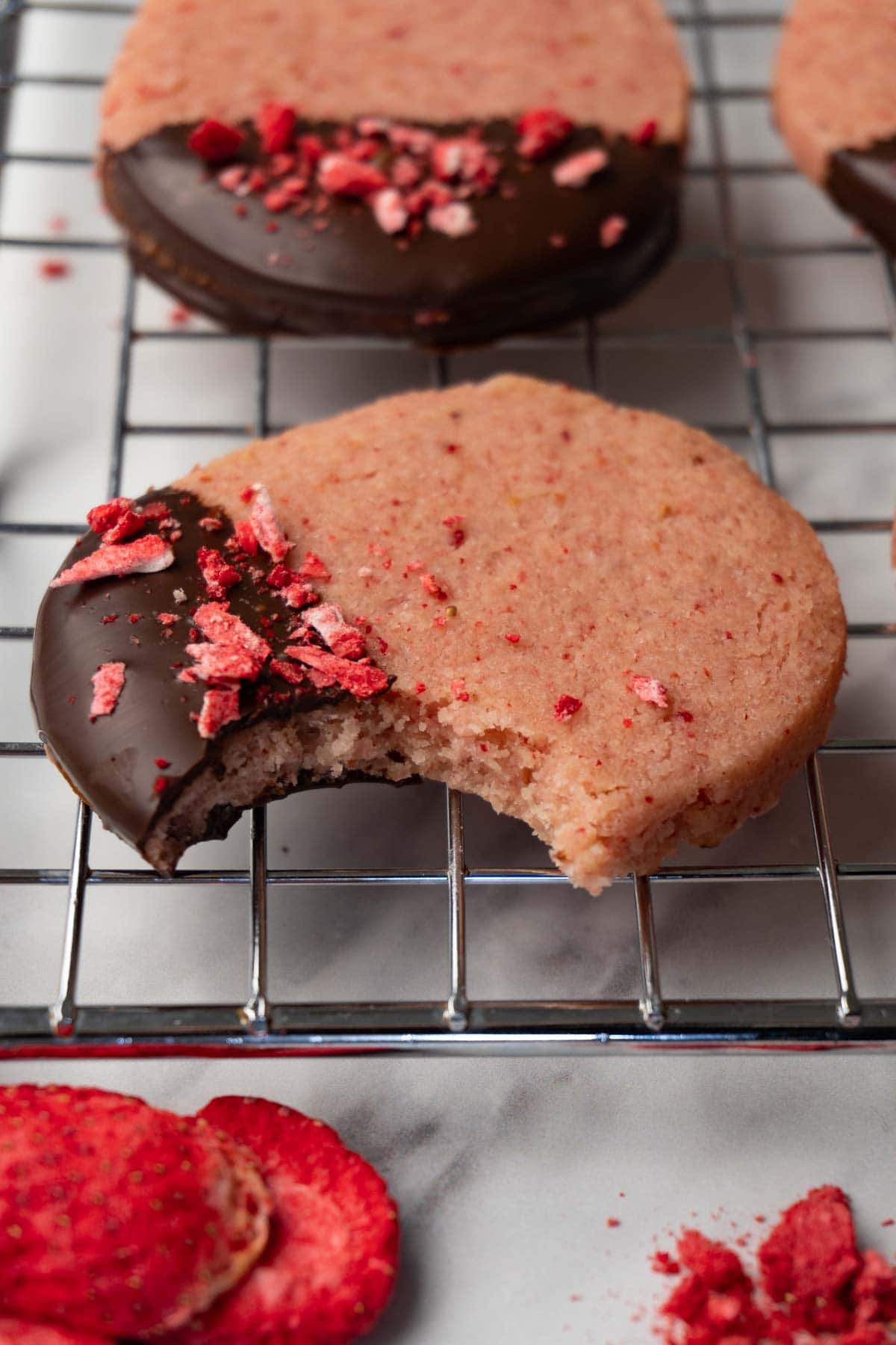 Halfway covered with dark chocolate strawberry shortbread cookies decorated with crushed freeze-dried strawberries are lying on a wire rack; one bite was taken from one of the cookies.