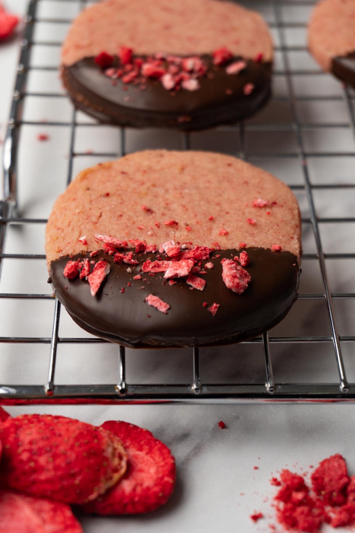 Halfway covered with dark chocolate strawberry shortbread cookies decorated with crushed freeze-dried strawberries are lying on a wire rack.
