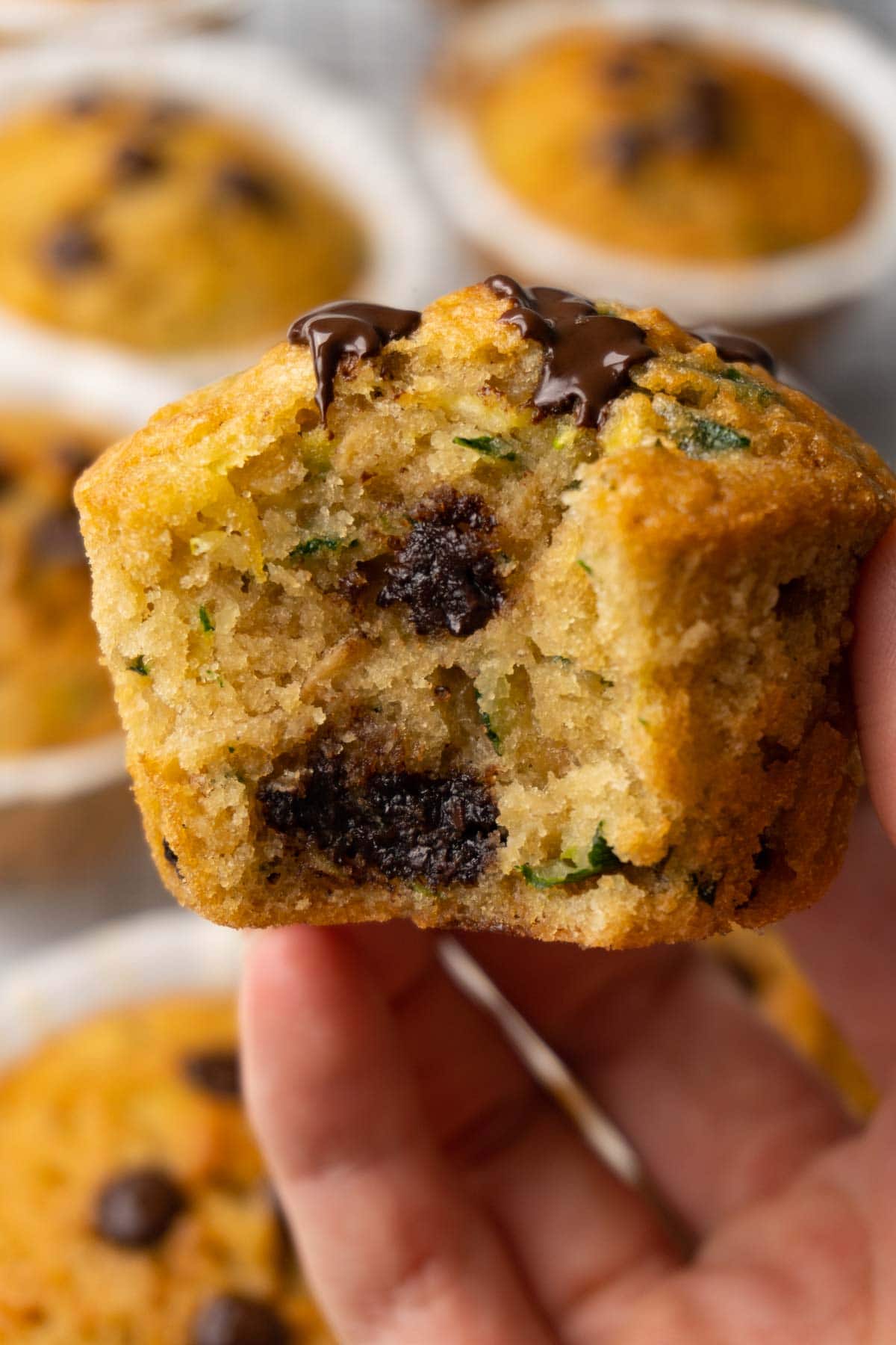 A hand is holding a muffin with chocolate chips and grated zucchini; one bite is taken.