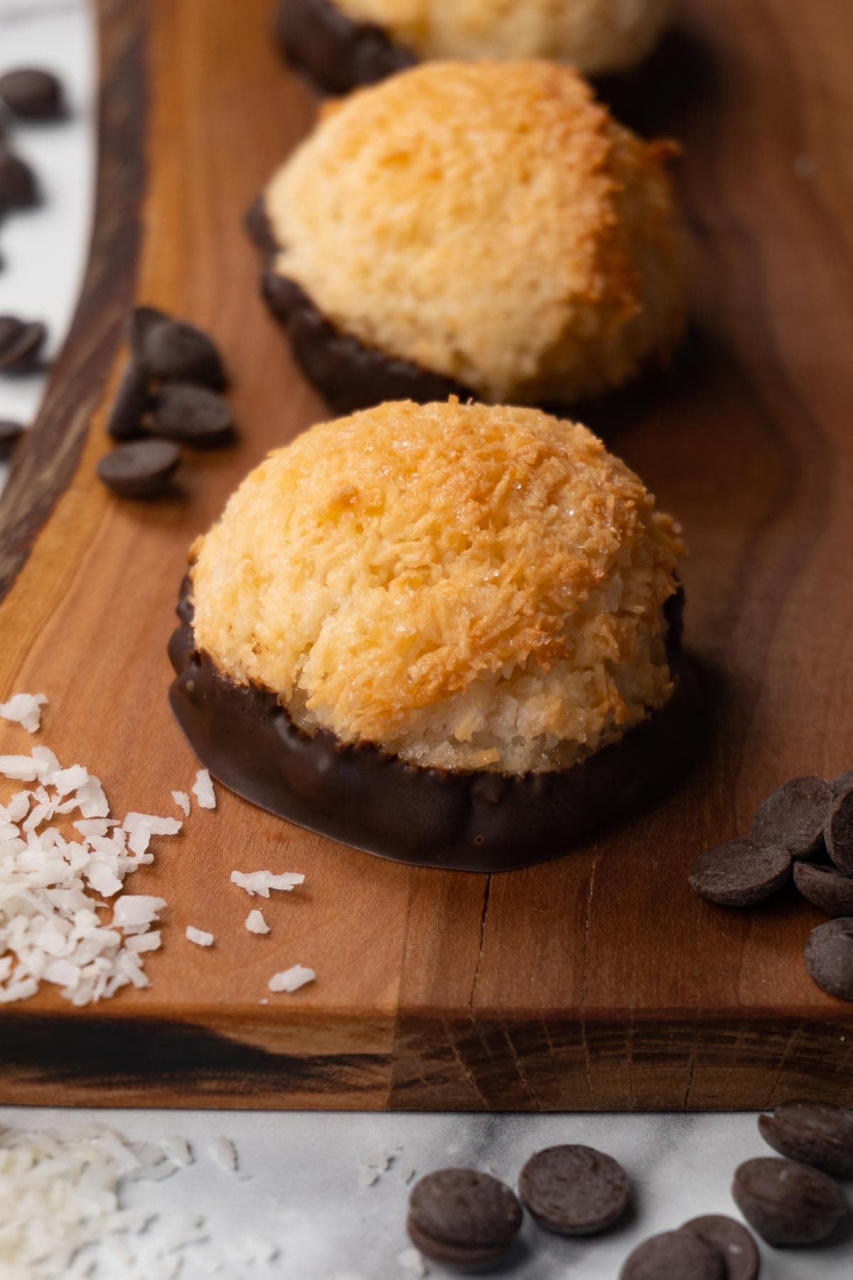 Coconut macaroons dipped in chocolate on a wooden board, coconut flakes, and chocolate chips are lying around.