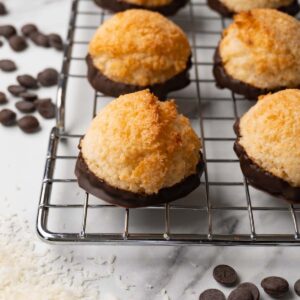 Macaroons dipped in chocolate on a wire rack, coconut flakes, and chocolate chips are lying around.