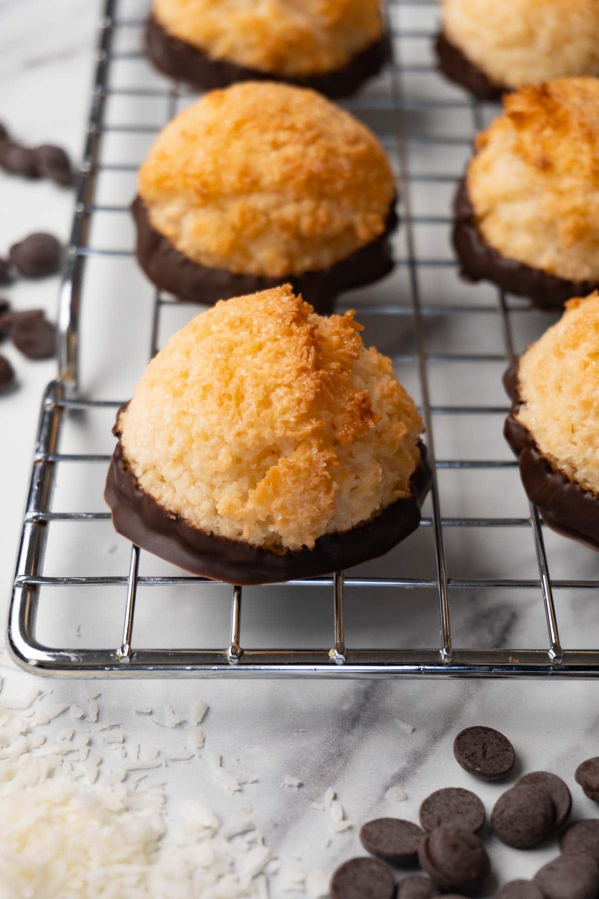 Macaroons dipped in chocolate on a wire rack, coconut flakes, and chocolate chips are lying around.