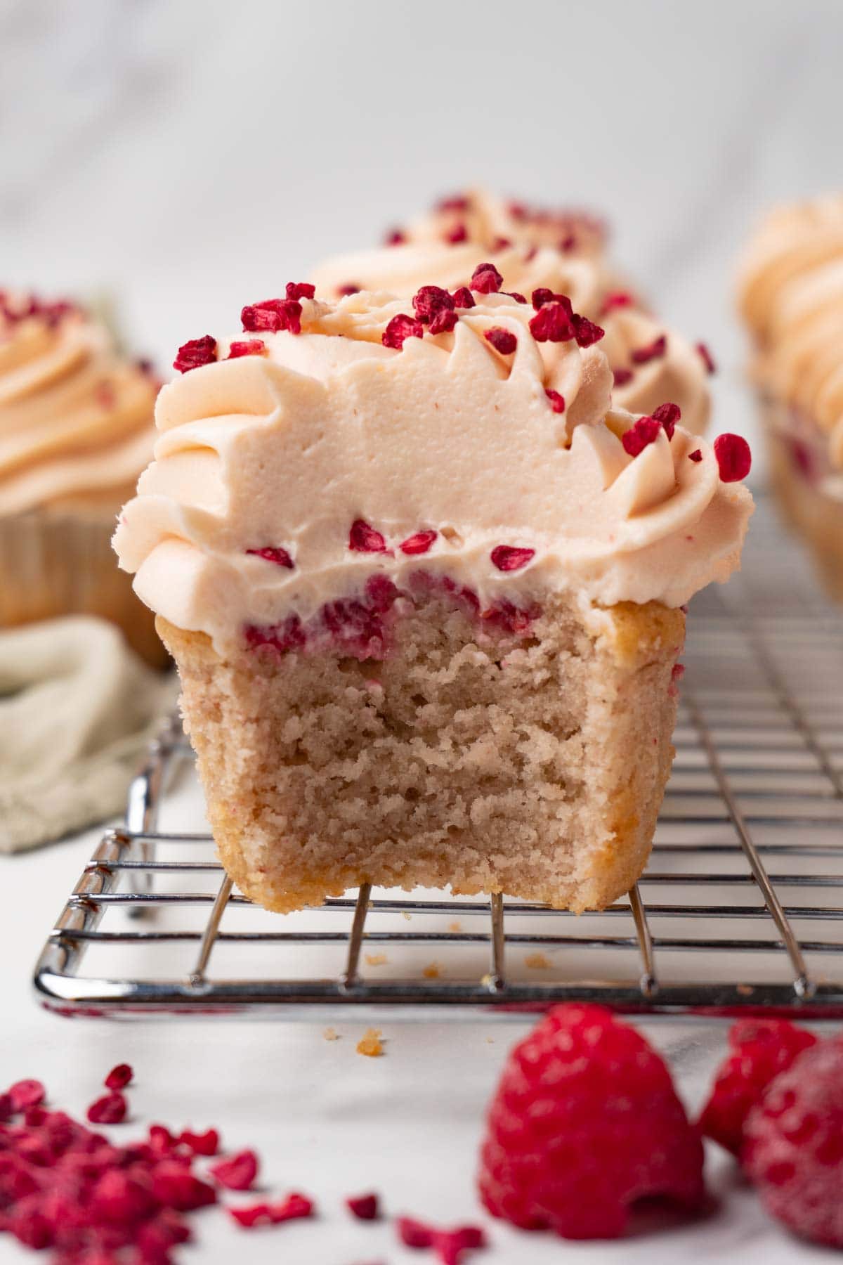 A cupcake with raspberries decorated with raspberry cream cheese frosting is standing on a wire rack. One bite was taken.